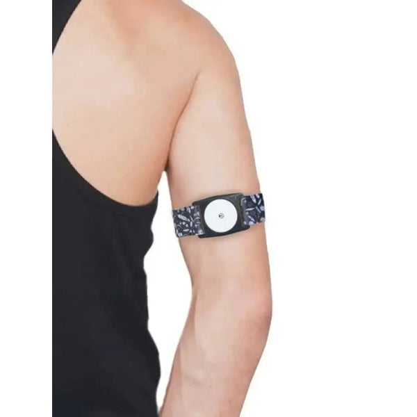A person wearing a Freestyle Libre 2 Armband with a patterned strap on their upper arm. The armband securely holds the sensor.